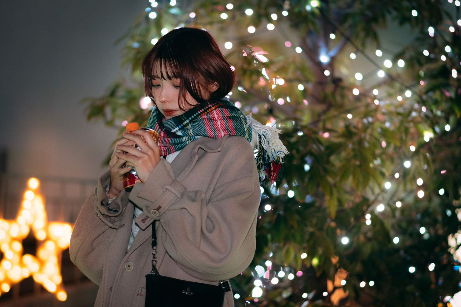 「彼の分の温かい飲み物で手を温めながらクリスマスツリーの前で待つ彼女」の写真［モデル：高尾実生］