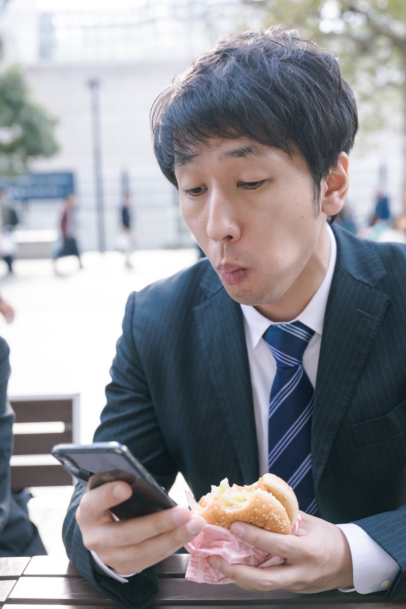 「ファーストフードを食べながらスマホをチェックする現代サラリーマン」の写真［モデル：大川竜弥］