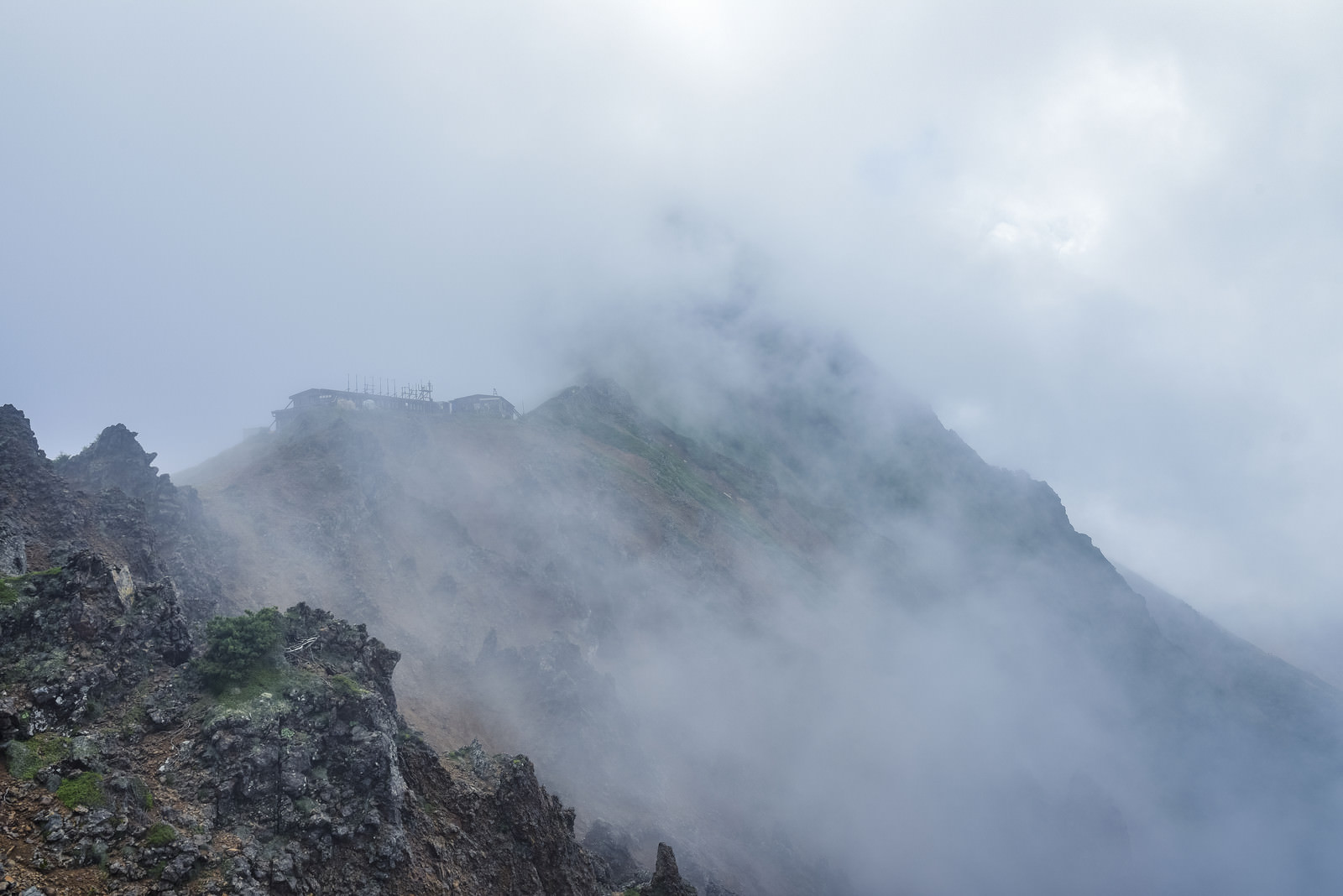 「雲の中にうっすらと姿を見せる赤岳と小屋 | フリー素材のぱくたそ」の写真