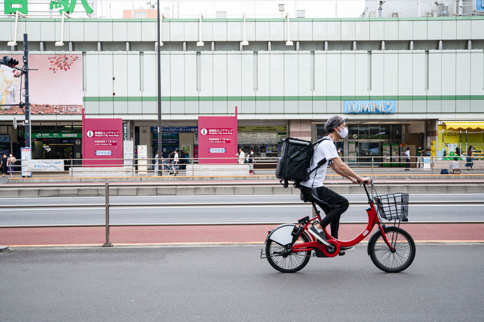 「新宿駅前を走行するフードデリバリーの配達員 | フリー素材のぱくたそ」の写真